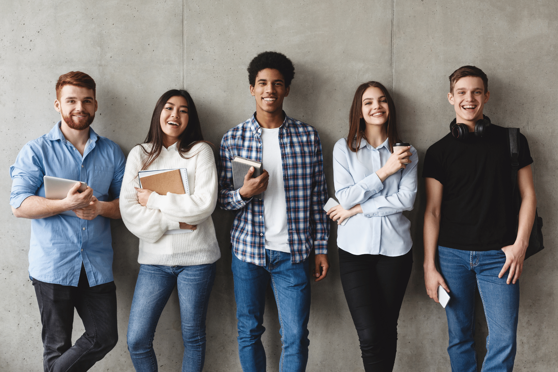 Group of smiling college students holding books and devices against a gray wall