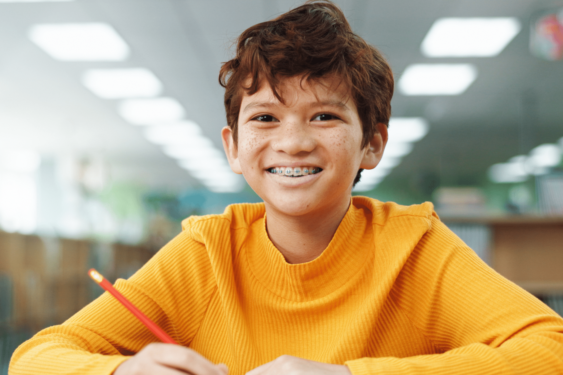 Smiling young boy wearing metal braces and holding a pencil in a classroom.